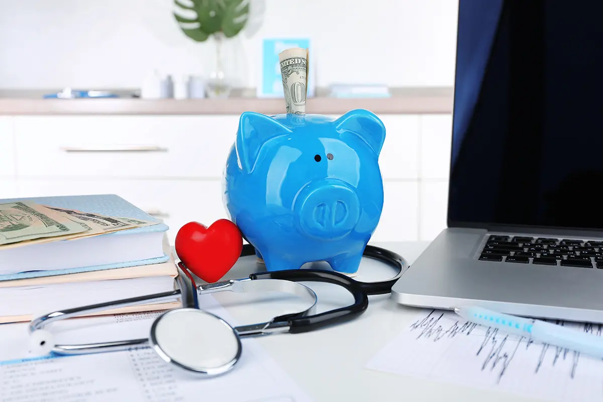 Blue piggy bank with dollar surrounded by stethoscope beside a laptop