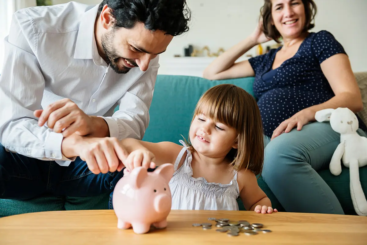 Little girl putting coins into piggy bank with father. Pregnant mother smiling in background.